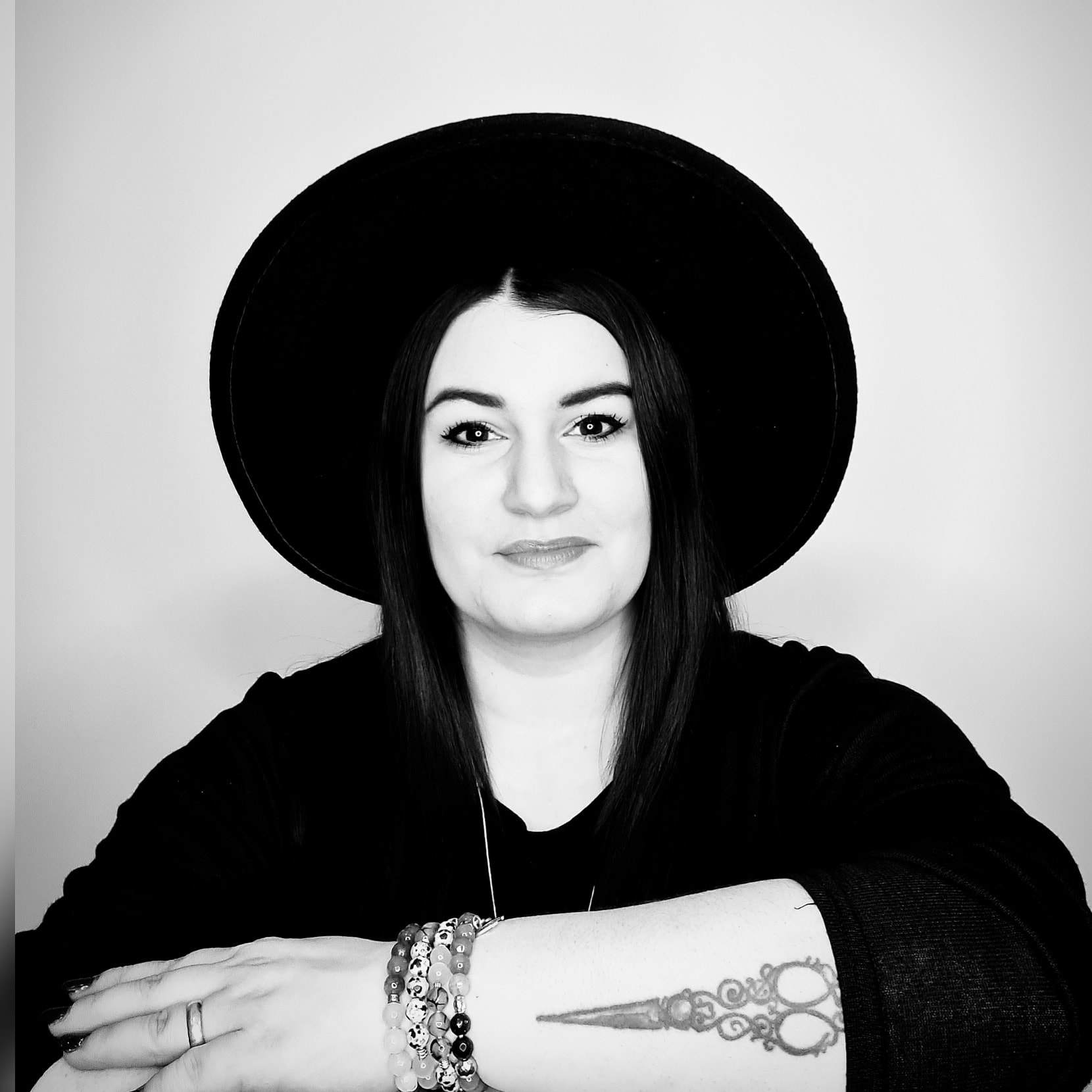 Hair Company UK The image labeled as 290345552_435017278634684_7480172814251602787_n showcases a black and white portrait of an individual with long hair, adorned in a wide-brimmed hat and a dark top. Standing confidently with arms crossed, the person displays bracelets and features a prominent scissors tattoo on their forearm, subtly reflecting their artistic training. A gentle smile graces their lips.