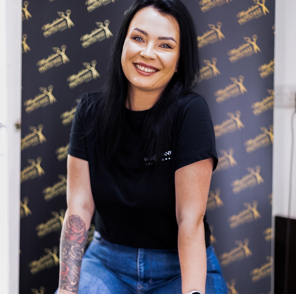 Hair Company UK A smiling woman with long dark hair and a rose tattoo on her forearm, wearing a black t-shirt and jeans, sits in front of the "456254527_1029278225865201_7047640073418679431_n" black and gold backdrop, exemplifying the refined artistry achieved through expert training.