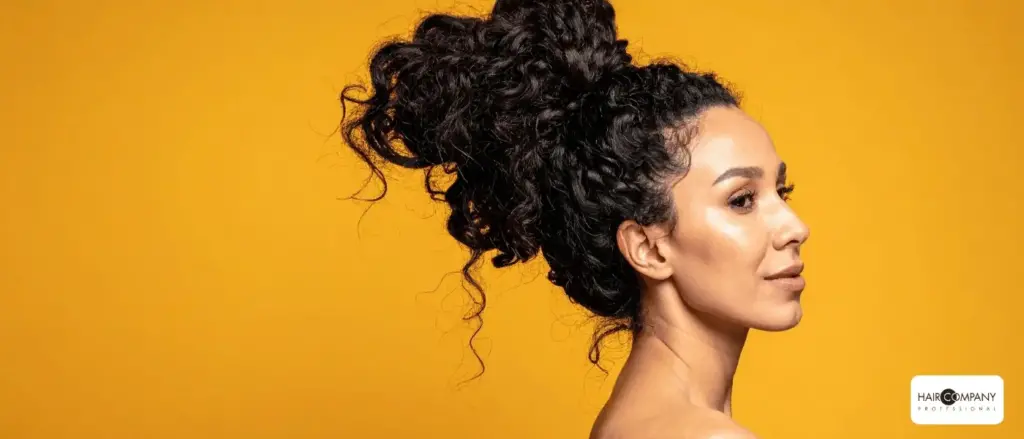 a woman with curly dark hair styled in a high bun poses against a vibrant yellow background, her glowing skin in profile. the "hair company uk distributor (2).opti" logo is displayed in the bottom right corner. hair company uk