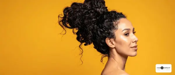 a woman with curly dark hair styled in a high bun poses against a vibrant yellow background, her glowing skin in profile. the "hair company uk distributor (2).opti" logo is displayed in the bottom right corner. hair company uk
